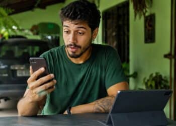 Homem olhando o celular à mesa, com um tablet à frente e um carro ao fundo, cena que representa as novidades do WhatsApp para o telefone, o tablet e o carro.