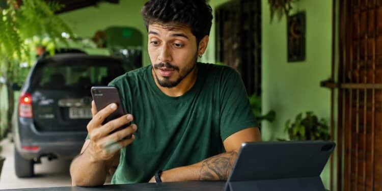 Homem olhando o celular à mesa, com um tablet à frente e um carro ao fundo, cena que representa as novidades do WhatsApp para o telefone, o tablet e o carro.