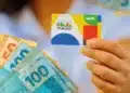 Pessoa segura cartão do Bolsa Família ao lado de cédulas de dinheiro, em referência a novo auxílio aprovado para beneficiários do programa.