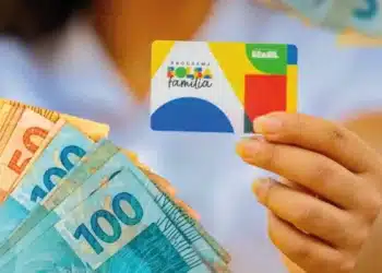Pessoa segura cartão do Bolsa Família ao lado de cédulas de dinheiro, em referência a novo auxílio aprovado para beneficiários do programa.