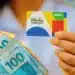 Pessoa segura cartão do Bolsa Família ao lado de cédulas de dinheiro, em referência a novo auxílio aprovado para beneficiários do programa.