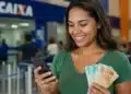 Mulher consulta pagamento pelo celular enquanto segura notas de real em agência bancária, representando o pagamento do Abono Salarial e da restituição do Imposto de Renda.