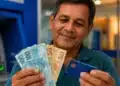 Homem segura notas de real e cartão bancário diante de um caixa eletrônico, em referência ao pagamento do 13º salário em novembro.