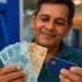 Homem segura notas de real e cartão bancário diante de um caixa eletrônico, em referência ao pagamento do 13º salário em novembro.