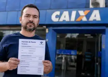 Contribuinte segura declaração do Imposto de Renda em frente à Caixa, em imagem que representa a espera pelos últimos lotes da restituição de 2026.
