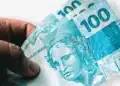 Mão segurando notas de 100 reais do Banco Central do Brasil, representando o pagamento do programa Pé-de-Meia em 1º de julho.