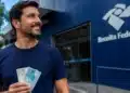 Homem segura cédulas de real em frente a uma unidade da Receita Federal, representando o pagamento de R$ 500 milhões a contribuintes.