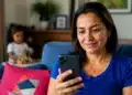 Mulher brasileira consultando smartphone em casa enquanto criança aparece ao fundo, representando o pagamento automatizado de pensão alimentícia pelo Pix