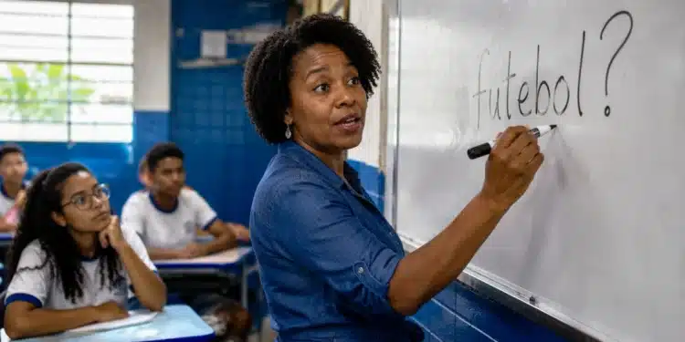 Professora escrevendo “futebol?” no quadro durante aula de português, representando a dúvida sobre o plural da palavra.