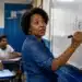 Professora escrevendo “futebol?” no quadro durante aula de português, representando a dúvida sobre o plural da palavra.