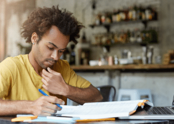 Estudante concentrado estudando edital de concurso público com notebook e apostilas.