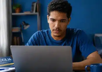 Jovem estudando pelo computador ao lado de um livro do Encceja, representando a dúvida sobre fazer a prova pela internet em 2026.