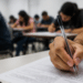 Mão segurando caneta durante prova escrita em sala de aula, representando concursos públicos com possíveis alterações de datas.