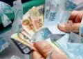 Cédulas de reais em mãos sobre imagem de dinheiro, representando a restituição do Imposto de Renda 2026, que libera lote regular e especial.