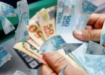 Cédulas de reais em mãos sobre imagem de dinheiro, representando a restituição do Imposto de Renda 2026, que libera lote regular e especial.