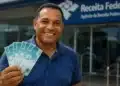 Homem segurando notas de real em frente à Receita Federal, representando o pagamento do 3º lote da restituição do Imposto de Renda em 2026