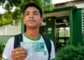 Estudante do ensino médio segurando nota de real em frente a uma escola pública, representando o pagamento do Pé-de-Meia em agosto.