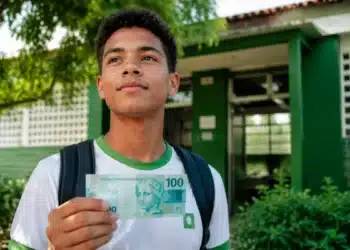 Estudante do ensino médio segurando nota de real em frente a uma escola pública, representando o pagamento do Pé-de-Meia em agosto.