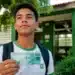 Estudante do ensino médio segurando nota de real em frente a uma escola pública, representando o pagamento do Pé-de-Meia em agosto.
