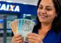 Mulher em agência da Caixa Econômica segura cédulas de real, simbolizando a liberação do saque-aniversário do FGTS em julho