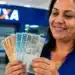 Mulher em agência da Caixa Econômica segura cédulas de real, simbolizando a liberação do saque-aniversário do FGTS em julho