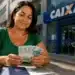 Mulher segurando notas de real em frente a uma agência da Caixa representando o saque de R$ 6.220 do FGTS