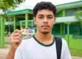 Jovem estudante brasileiro segurando o cartão do programa Pé-de-Meia em frente a uma escola pública.