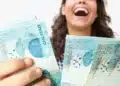 Mulher segura notas de real, representando o pagamento do terceiro lote da restituição do Imposto de Renda a 2,7 milhões de contribuintes.