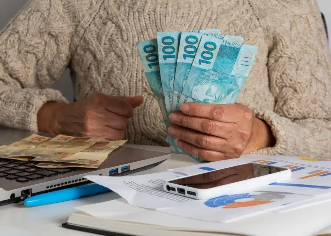 Formas de usar o FGTS para reduzir o saldo do financiamento Pessoa segurando notas de real ao lado de documentos e computador, representando o uso do FGTS para amortização ou quitação de financiamento imobiliário.