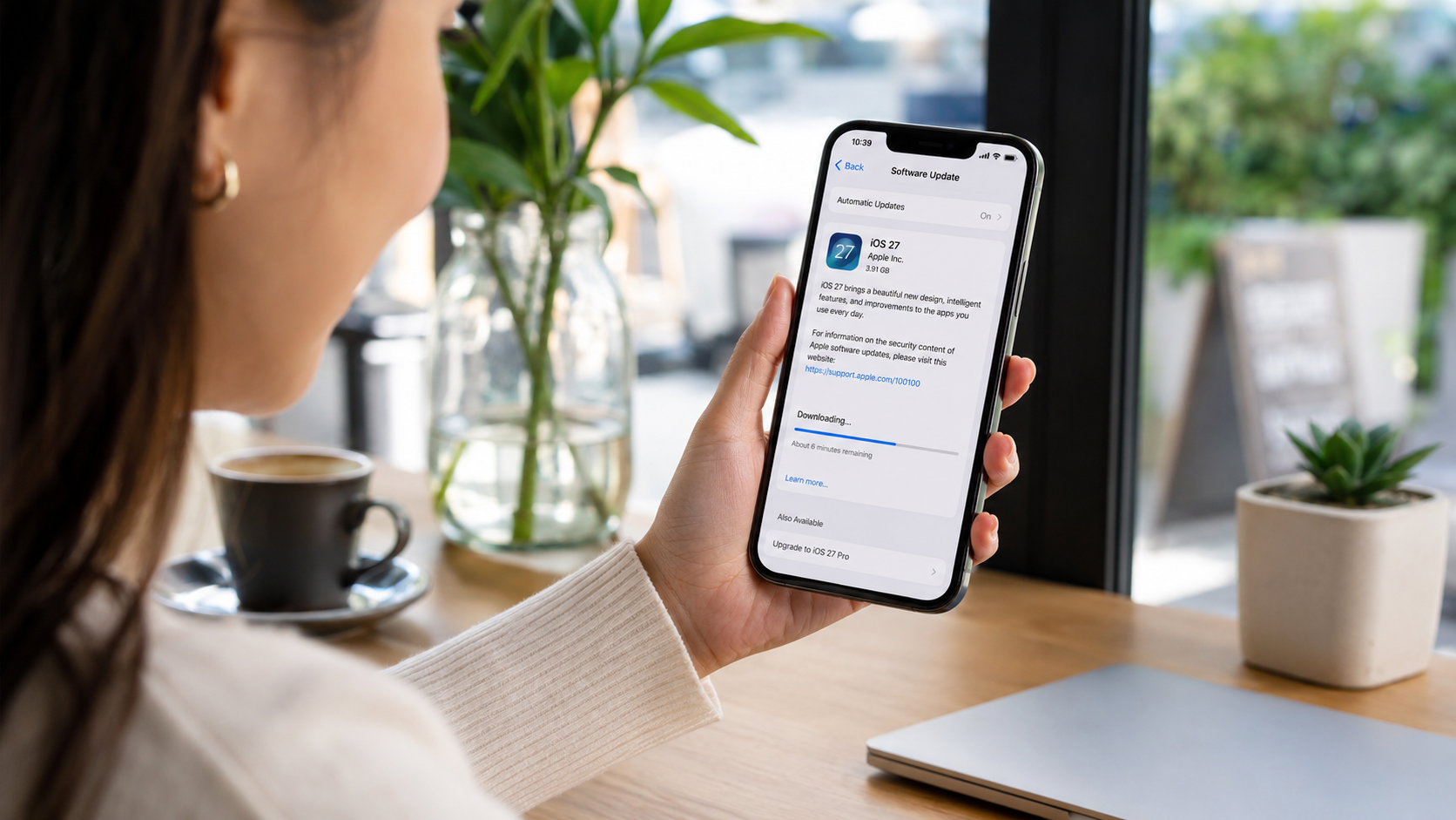 Mão feminina segura iPhone exibindo tela de download do iOS 27, com barra de progresso azul, em ambiente de café.