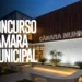 Fachada iluminada de Câmara Municipal com letreiro e anúncio de concurso público em destaque.