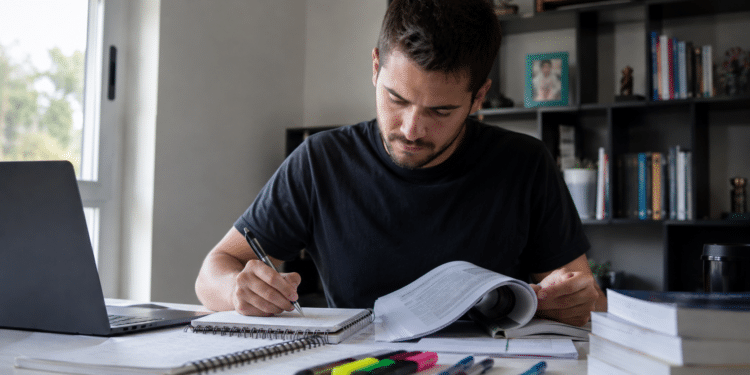 Homem jovem escrevendo em caderno enquanto consulta livro aberto, laptop e materiais de estudo sobre mesa.