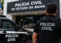 Policial civil de costas em frente à sede da Polícia Civil da Bahia, viatura ao lado.
