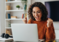 Jovem comemorando conquista ao computador ambiente doméstico alegre.