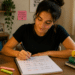 Jovem mulher escreve em caderno de gramática, rodeada de livros de português e post-its motivacionais.