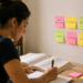 Mulher escreve em caderno ao lado de livros abertos e post-its de Direito Administrativo na parede.