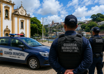 Dois guardas municipais de colete observam praça com viatura em frente à igreja colonial em Valença-BA.