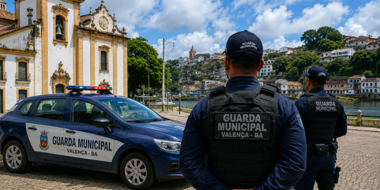 Dois guardas municipais de colete observam praça com viatura em frente à igreja colonial em Valença-BA.