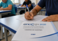 Pessoa escreve em folha ao lado de caderno Encceja 2026 em sala com outras pessoas ao fundo.