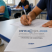 Pessoa escreve em folha ao lado de caderno Encceja 2026 em sala com outras pessoas ao fundo.