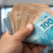 Pessoa segura notas de 50 e 100 reais em ambiente doméstico.