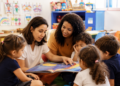 Duas mulheres lendo livro ilustrado para quatro crianças sentadas em roda em sala decorada com desenhos.