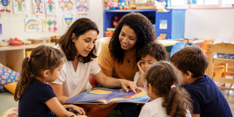 Duas mulheres lendo livro ilustrado para quatro crianças sentadas em roda em sala decorada com desenhos.