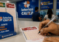 Homem escreve com caneta presa por corrente em volante da Loteca, expositores de apostas da Loterias Caixa ao fundo.
