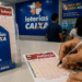 Homem escreve com caneta presa por corrente em volante da Loteca, expositores de apostas da Loterias Caixa ao fundo.
