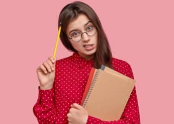 Imagem ilustrativa de jovem de óculos e camisa vermelha de bolinhas segurando um caderno espiral e um lápis junto à cabeça, com expressão de dúvida, remetendo à insegurança na hora de grafar a palavra corretamente