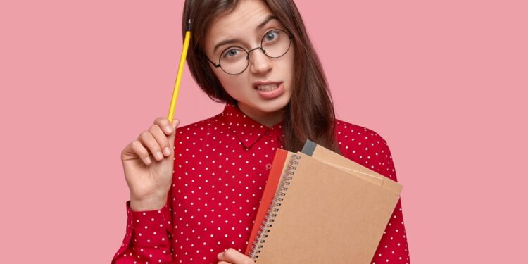 Imagem ilustrativa de jovem de óculos e camisa vermelha de bolinhas segurando um caderno espiral e um lápis junto à cabeça, com expressão de dúvida, remetendo à insegurança na hora de grafar a palavra corretamente