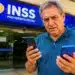 Homem idoso confere celulares em frente a uma agência do INSS, representando a manutenção do acesso ao Meu INSS após a troca de aparelho.