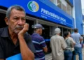 Homem aguarda atendimento diante de agência do INSS enquanto fila de beneficiários se estende pela calçada