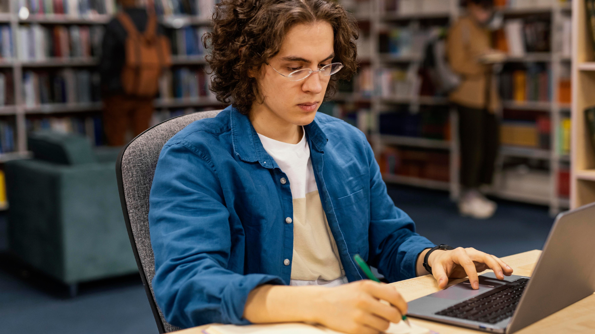 Estudante estudando em biblioteca com notebook e materiais de preparação para concurso público.
